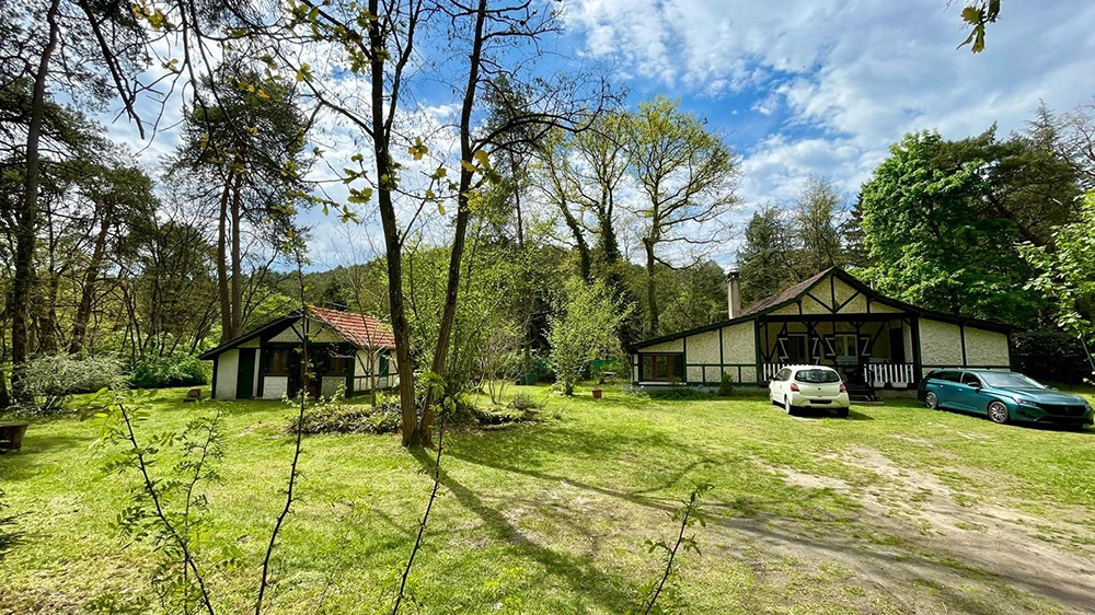 Global outdoor view of the terrain from when you arrive. Terrain is forest like with trees. On the left, apartment, on the right, house. Two cars are parked in front of the house.